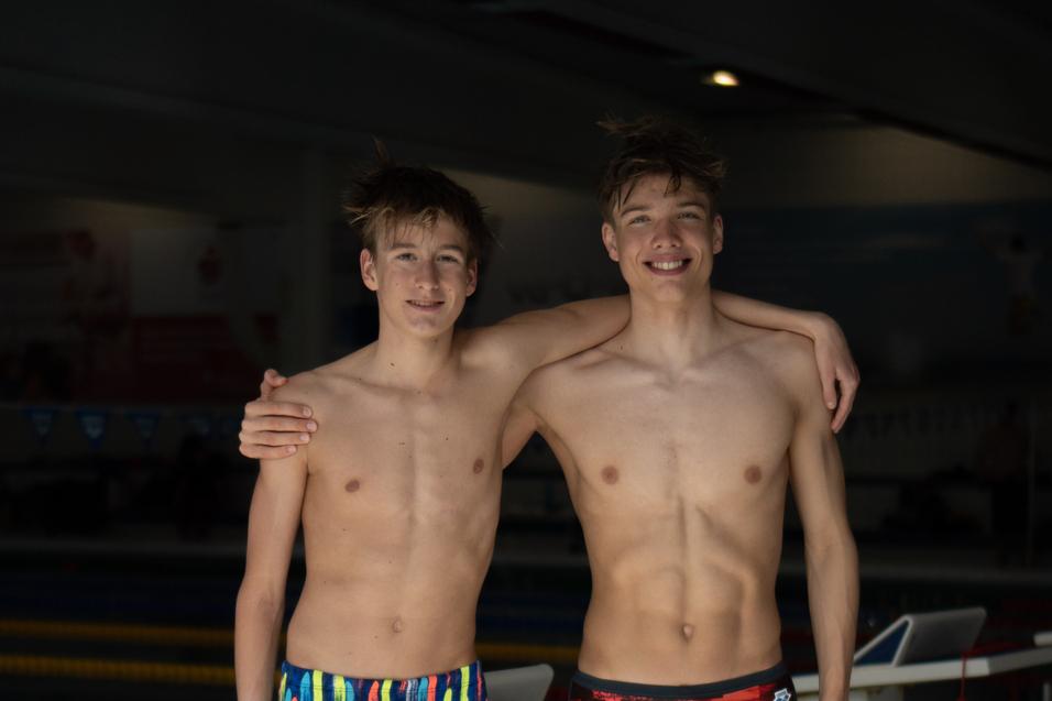 Zwei junge Schwimmer stehen am Beckenrand, einer in bunten Shorts, der andere in rot-schwarzen Badehosen.