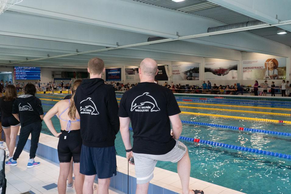 Zuschauer stehen am Beckenrand eines Schwimmwettkampfs in einer Halle mit bunten Bahnen.