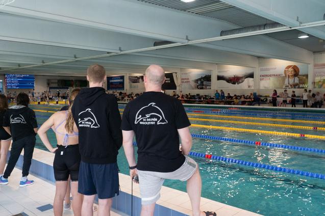 Zuschauer stehen am Beckenrand eines Schwimmwettkampfs in einer Halle mit bunten Bahnen.