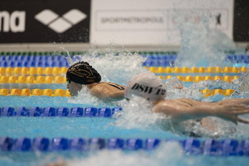 Zwei Schwimmerinnen im Wettkampf, eine mit schwarzer Badekappe und die andere mit weißer Badekappe, im Wasser.