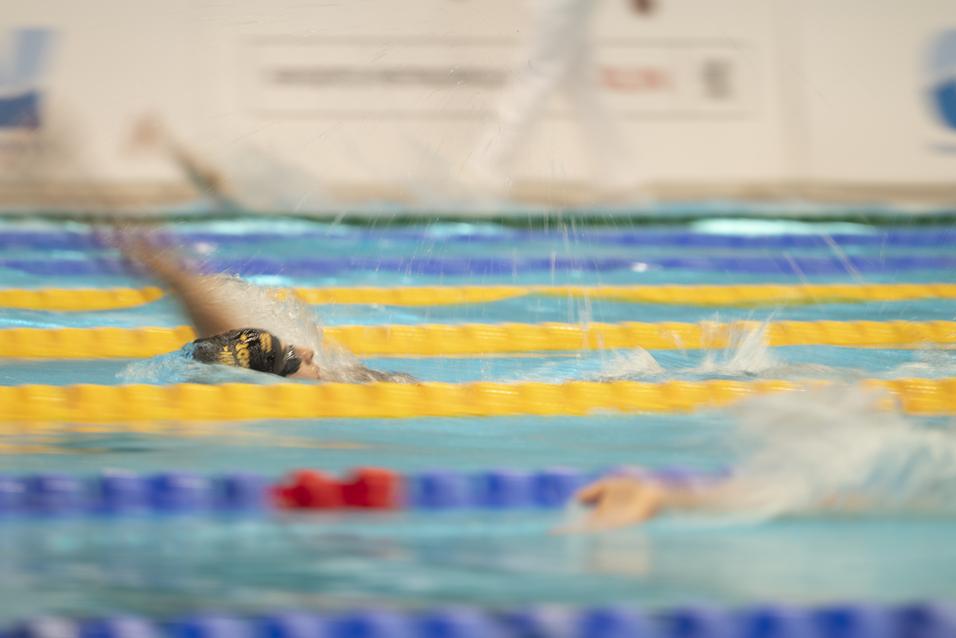 Schwimmerin im Wasser, während sie einen Wettkampf auf einer bunten Schwimmbahn bestreitet. Spritzwasser ist sichtbar.
