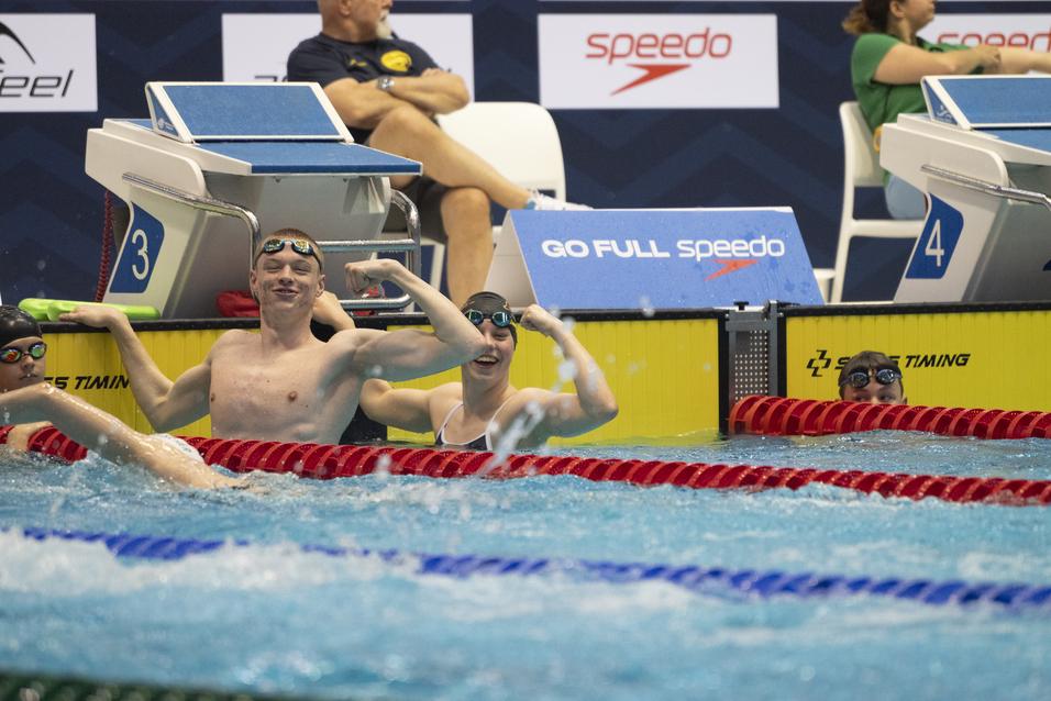 Junge Schwimmer posieren lächelnd am Beckenrand, während sie mit einem schwimmenden Mädchen interagieren.