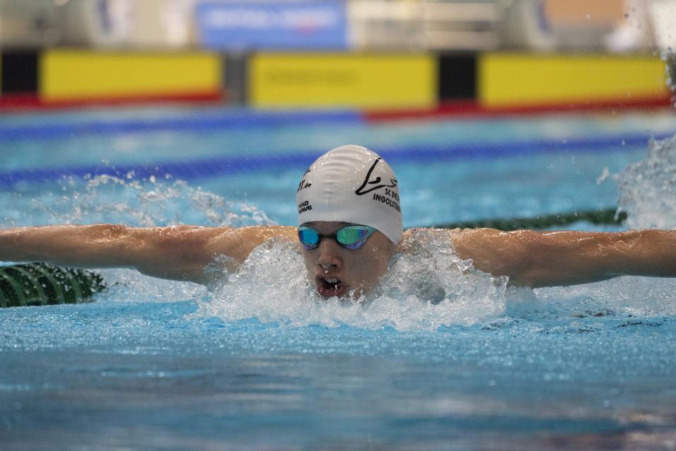 Schwimmer in Wettkampfkleidung führt einen Schmetterlingsschwimmstil im Pool aus, spritzendes Wasser um ihn herum.