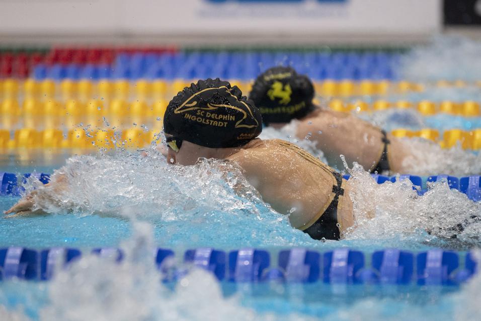 Zwei Schwimmer in schwarzen Kappen schwimmen im Wettkampfstil. Spritzendes Wasser in einem Pool mit bunten Bahnmarkierungen.