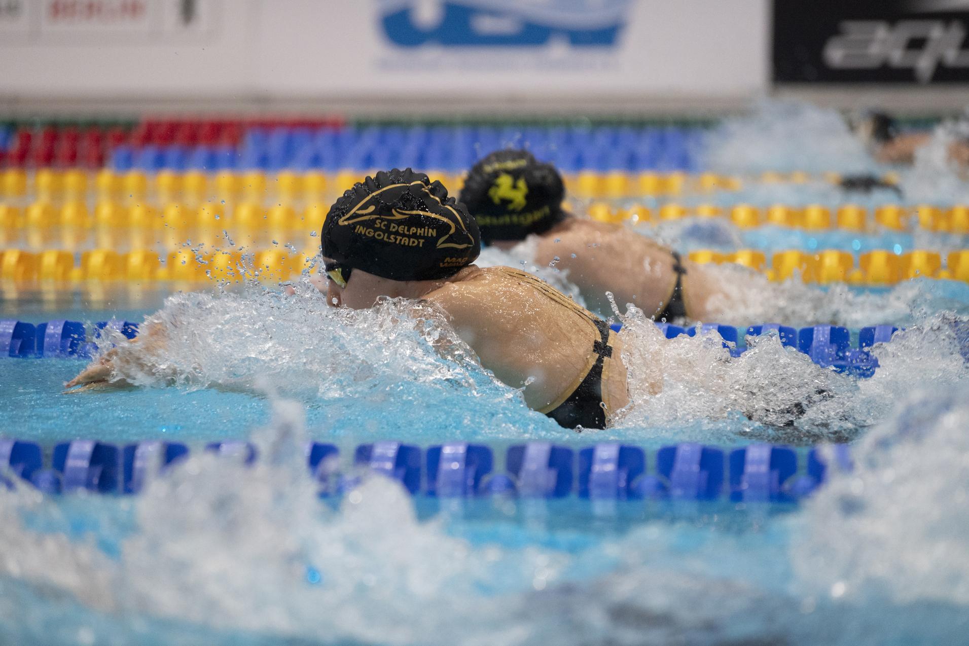 Zwei Schwimmer in schwarzen Kappen schwimmen im Wettkampfstil. Spritzendes Wasser in einem Pool mit bunten Bahnmarkierungen.
