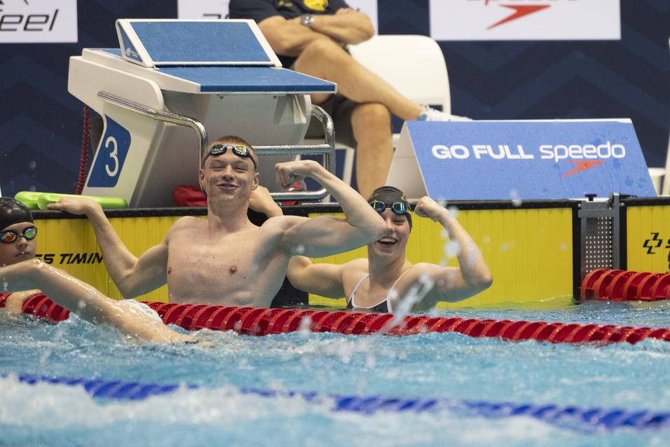 Junge Schwimmer posieren lächelnd am Beckenrand, während sie mit einem schwimmenden Mädchen interagieren.