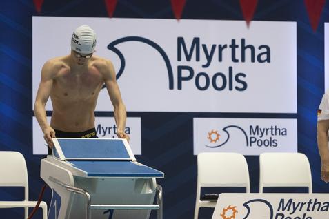 Schwimmer in Badehose mit Schwimmhaube beugt sich über einen Startblock vor einem Myrtha Pools Banner.