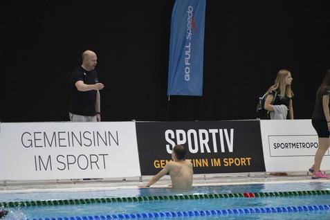 Ein Trainer steht am Beckenrand und gibt Anweisungen an einen Schwimmer, während Zuschauer im Hintergrund stehen.