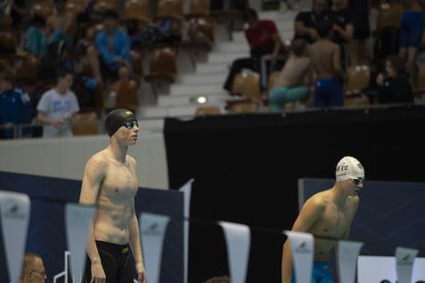 Zwei Schwimmer in Wettkampfbekleidung stehen am Beckenrand, bereit für den Start, mit Zuschauern im Hintergrund.