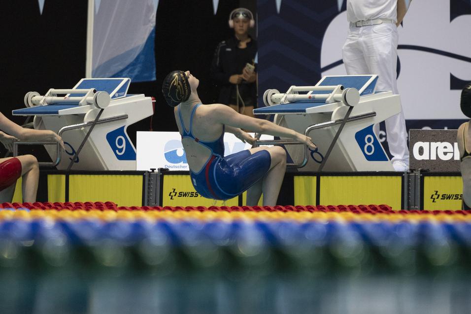 Zwei Schwimmerinnen in Startposition an Wettkampfblöcken, im Hintergrund ein Offizieller und eine weitere Person.