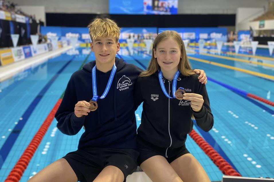 Zwei junge Schwimmer mit Medaillen sitzen am Beckenrand, im Hintergrund ist die Schwimmhalle zu sehen.