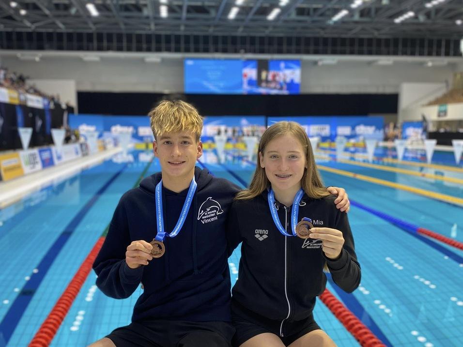 Zwei junge Schwimmer mit Medaillen sitzen am Beckenrand, im Hintergrund ist die Schwimmhalle zu sehen.
