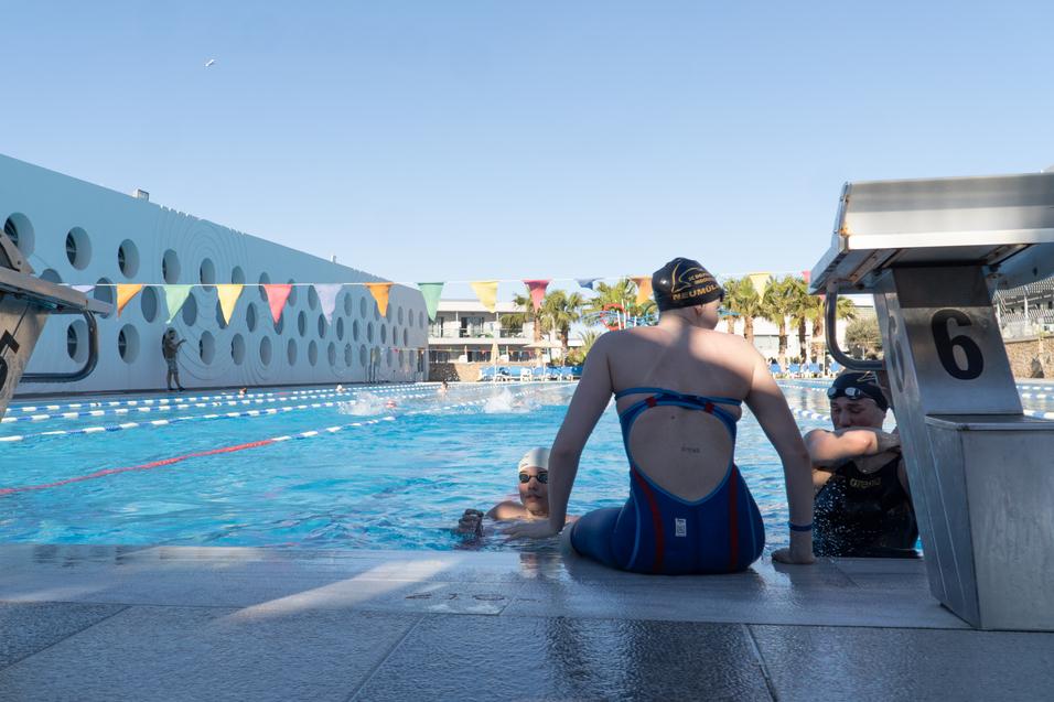 Drei Schwimmerinnen am Beckenrand eines Schwimmbads, mit Startblöcken und bunten Fahnen im Hintergrund.