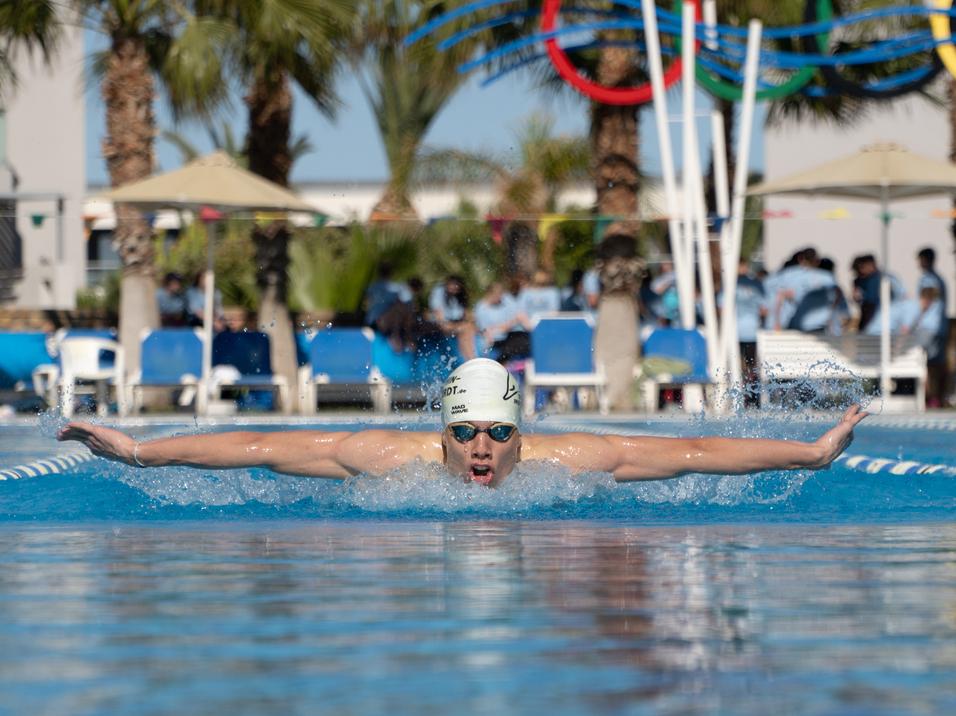 Schwimmer in einem weißen Badekappe zieht beim Schmetterlingsstil durch das Wasser einer blauen Schwimmhalle.
