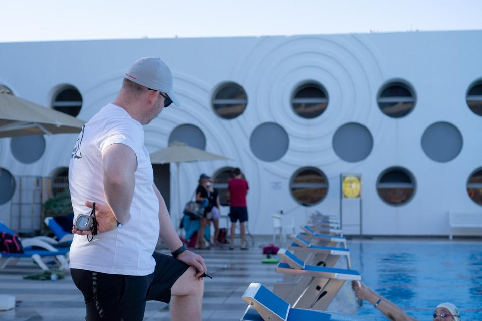 Ein Schwimmaufseher in T-Shirt und Hut beobachtet Schwimmer neben einer blauen Poolkante und sonnigen Liegen.