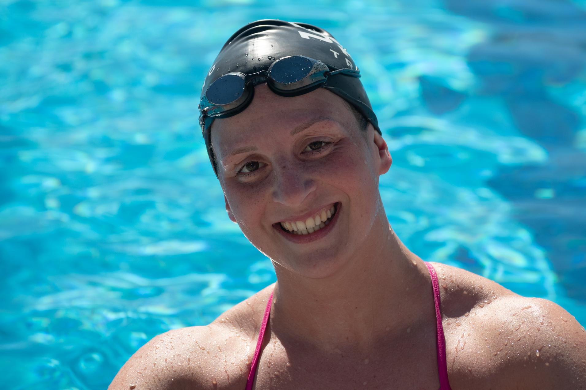 Lächelnde Schwimmerin in pinkem Badeanzug und Schwimmhaube posiert vor blauem Wasser eines Schwimmbeckens.