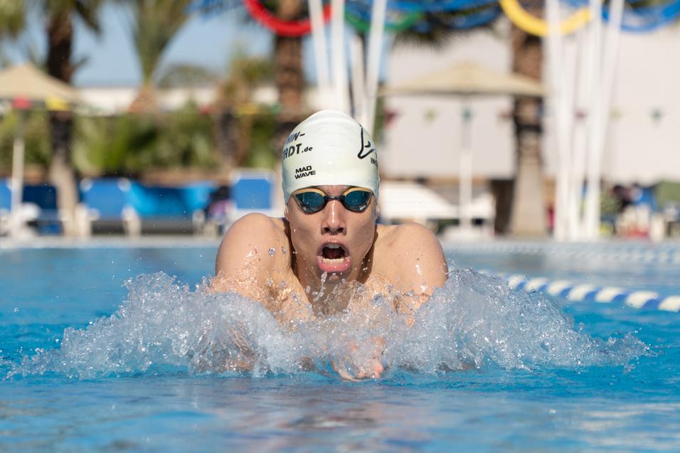 Schwimmer führt einen Kraulzug im Pool aus, spritzt Wasser und trägt eine Schwimmhaube sowie Schwimmbrille.