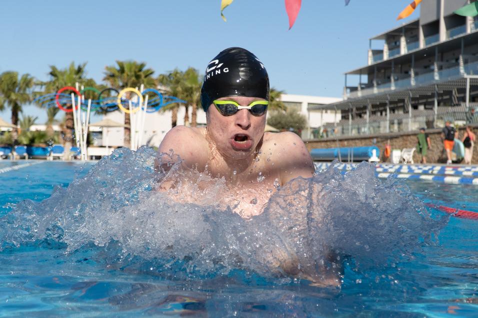 Schwimmer in dunkler Badekappe und grünen Schwimmbrille sprintet durch das Wasser, spritzt dabei Wasser auf.