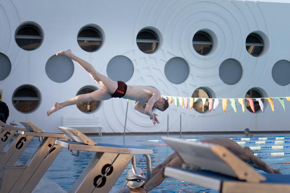 Sprinter springt kopfüber vom Startblock in ein Schwimmbecken mit bunten Wimpeln und runder Architektur im Hintergrund.